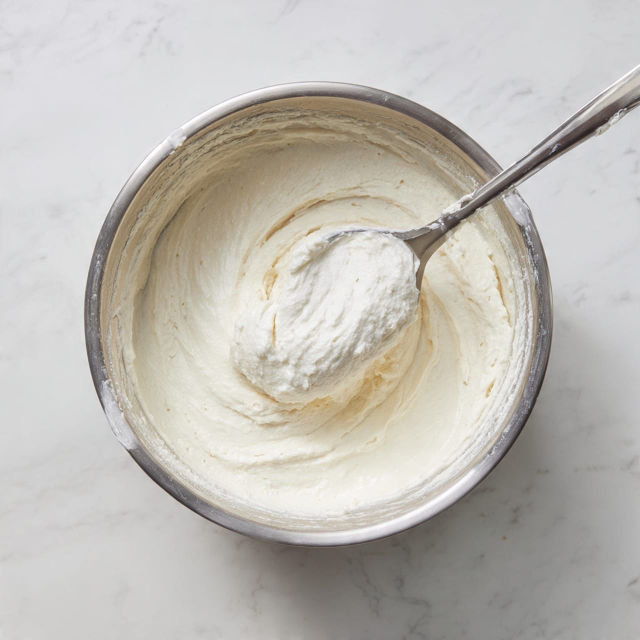 A close-up of a cupcake sliced in half showing two layers: a light golden soft cake base and a thick top layer of creamy white frosting swirled smoothly with a bit of texture. Colorful small round sprinkles in red, green, blue, pink, and yellow are scattered on the frosting. The cupcake sits in a white cupcake liner on a white marbled surface. Other whole cupcakes with the same frosting and sprinkles are blurred out in the background. photo taken with an iphone --ar 4:5 --v 7