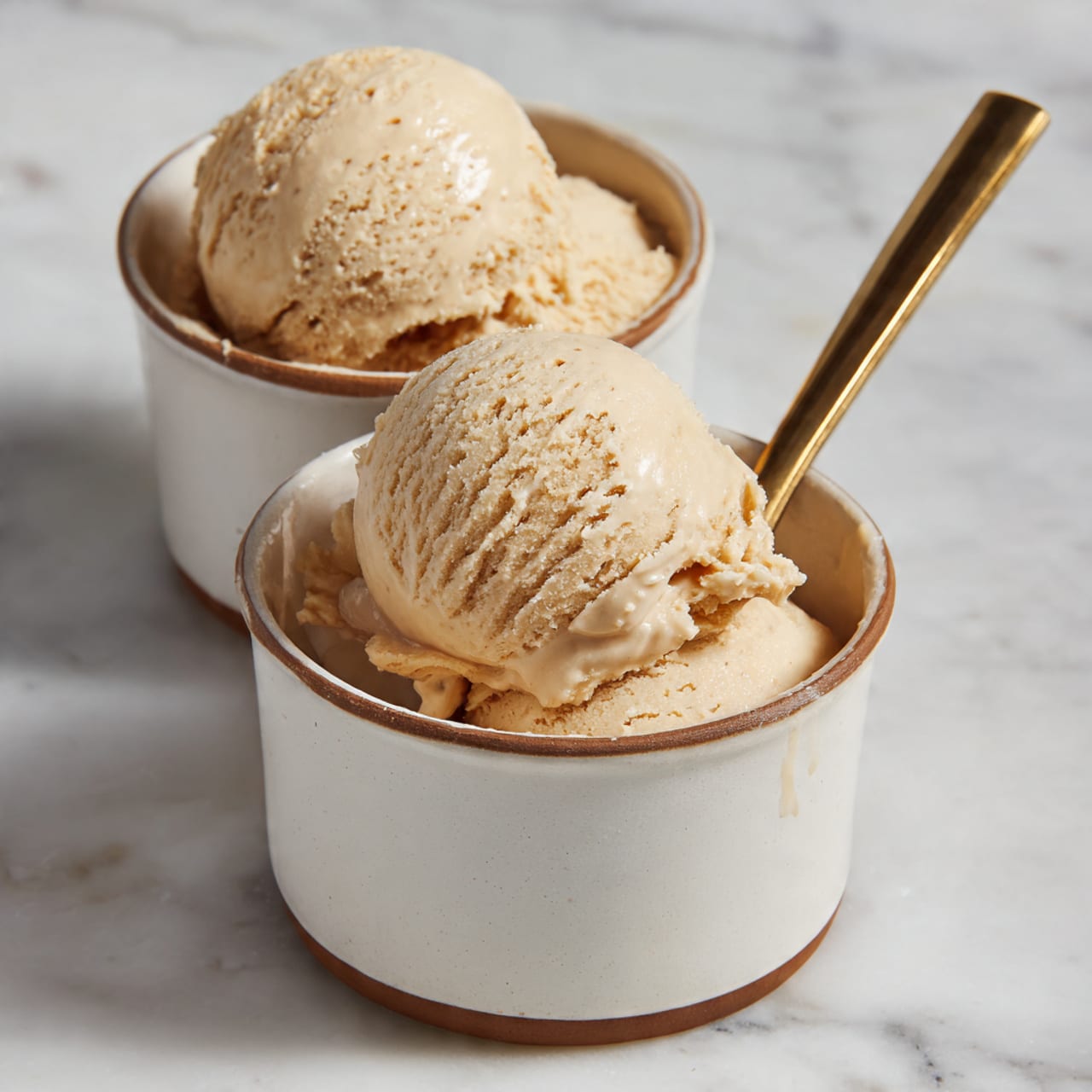 Two scoops of light brown ice cream with a smooth and creamy texture are placed inside a white cup with a brown rim. A thin golden spoon is inside the cup, resting against the ice cream. Behind this cup, there is another identical white cup with a brown rim, also filled with two scoops of the same ice cream. Both cups sit on a white marbled surface. The ice cream looks soft with small ridges and swirls from scooping. photo taken with an iphone --ar 4:5 --v 7