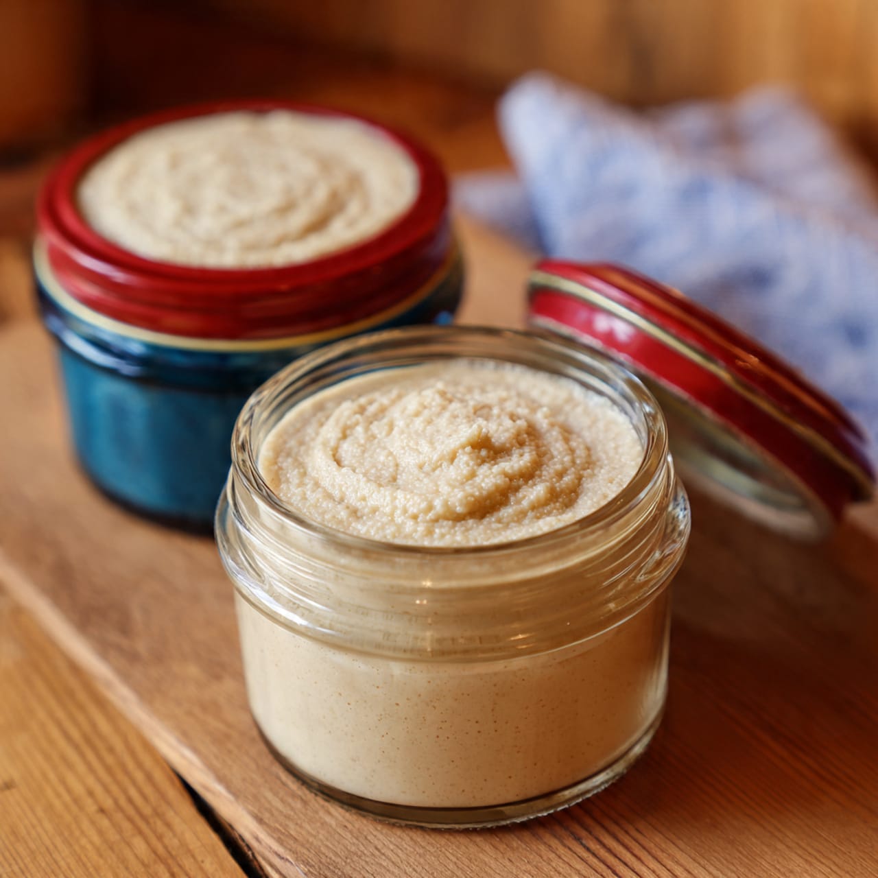 Two glass jars with metal clasps are filled with a creamy, light beige mixture, each jar showing a smooth texture with slight bubbles on top. The jars are placed side by side on a wooden board with visible grain and warm tones. The background is softly blurred, emphasizing the jars in the front. The surface beneath the board is a white marbled texture. photo taken with an iphone --ar 4:5 --v 7