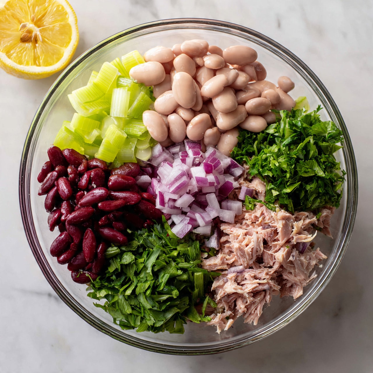 A clear glass bowl sits on a white marbled surface, holding five distinct layers of ingredients arranged in separate sections. One section has light pink beans with a smooth texture, next to a layer of pale green diced celery. Beside that is a small pile of bright green fresh parsley leaves. There are small pieces of purple-red chopped onions in the center, and a section with shredded light brown tuna. In the top left corner of the image, there is a lemon half with visible seeds. photo taken with an iphone --ar 4:5 --v 7