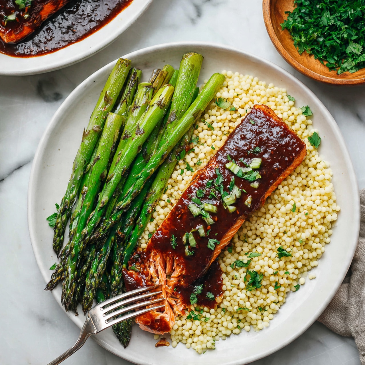 The image shows a white plate with three layers: the bottom layer is small, pale yellow couscous grains spread evenly across the plate; on top of the couscous is a dark brown grilled fish fillet with a shiny, slightly sticky glaze and small bits of green garnish; finally, five bright green grilled asparagus spears rest diagonally on the fish. The background features a white marbled surface with a white bowl of fish and a small white bowl filled with chopped green onions nearby. Photo taken with an iphone --ar 4:5 --v 7