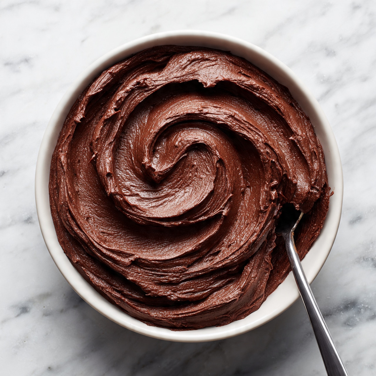 The image shows a single-layer round chocolate cake with a thick layer of smooth, rich, dark brown chocolate frosting spread evenly on top. The frosting has visible swirls and soft peaks creating texture across the surface. The cake itself is a deep, dark brown color, appearing moist and dense. It is placed on a white plate set on a white marbled surface. Photo taken with an iphone --ar 4:5 --v 7