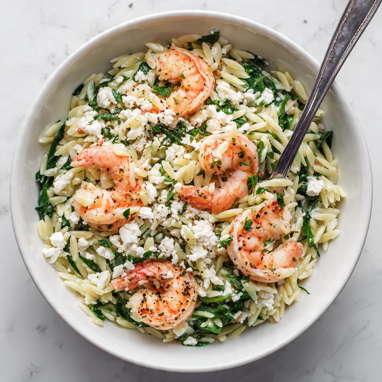 A white bowl filled with a shrimp and orzo salad on a white marbled surface. The dish has three main layers: the bottom layer is white orzo pasta with a slightly glossy texture, mixed evenly with bright green pieces of fresh spinach and parsley. The middle layer includes blotches of crumbled white feta cheese scattered across. On top are several pink-orange cooked shrimp, slightly glossy and seasoned with black pepper. A silver fork holds a bite, showing layers of shrimp, orzo, spinach, and feta. Photo taken with an iphone --ar 4:5 --v 7