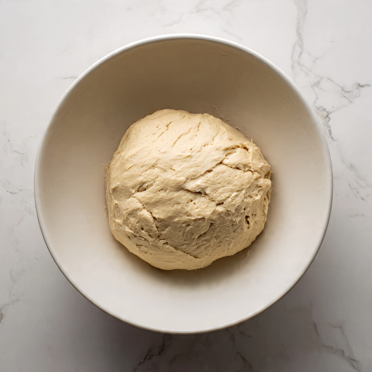 The image shows a close-up of a soft round bread roll with a light brown crispy top layer and a fluffy white inside layer visible where a bite has been taken, placed on a white plate. There are five more similar rolls in the background on white plates, all sitting on a white marbled surface. The bread rolls have a smooth golden-brown texture on top and a slightly darker but thin base layer, creating a dome shape. The focus is mainly on the bread roll in the front, while the others are softly blurred behind it photo taken with an iphone --ar 4:5 --v 7