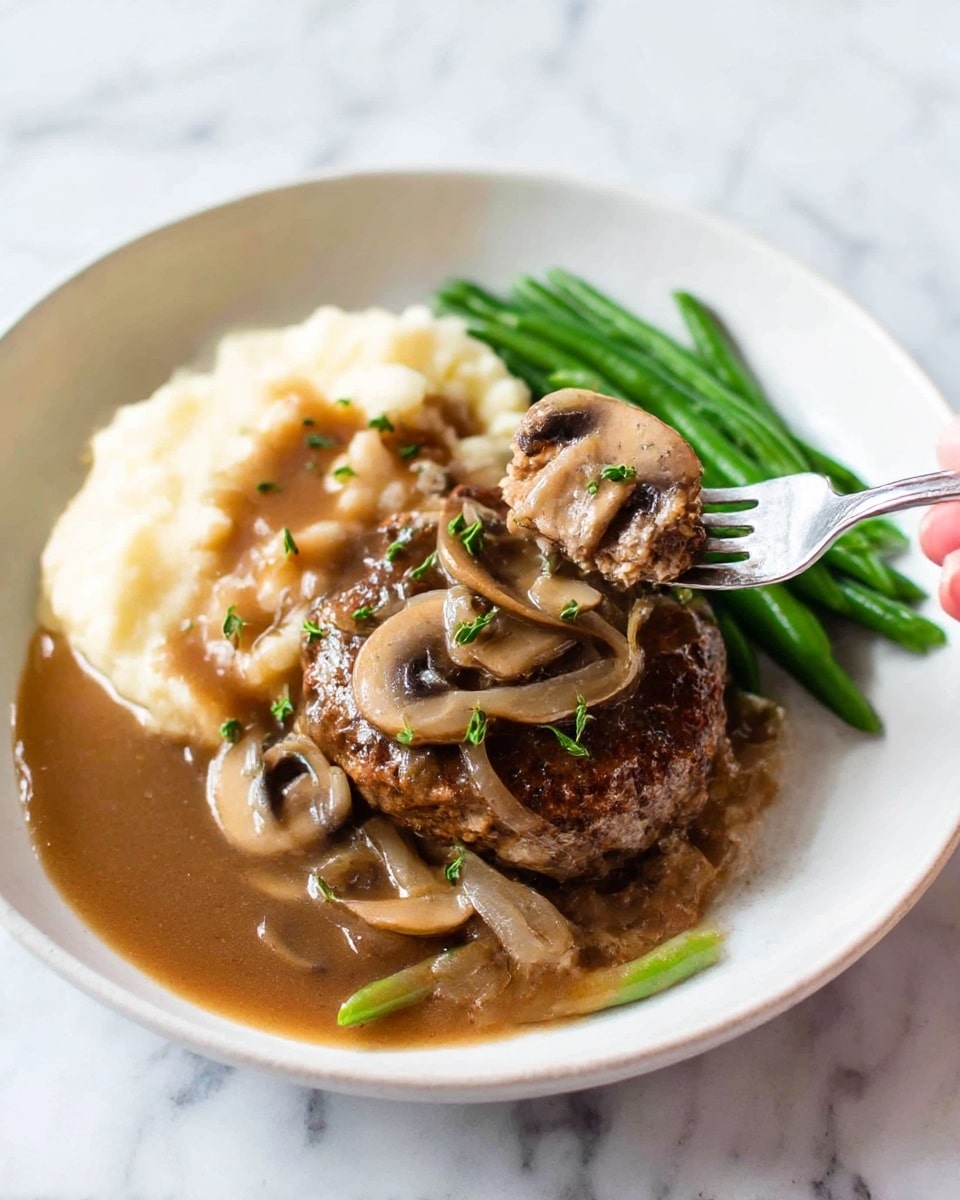 A close-up view of a browned meat patty in the center, covered with light brown mushroom sauce that has a smooth texture and visible mushroom slices. The sauce surrounds the patty and fills the black pan, with scattered small green herb bits on the sauce and mushrooms. A silver spoon is shown scooping up sauce and mushrooms from the right side of the pan. The scene is set on a white marbled surface. photo taken with an iphone --ar 4:5 --v 7