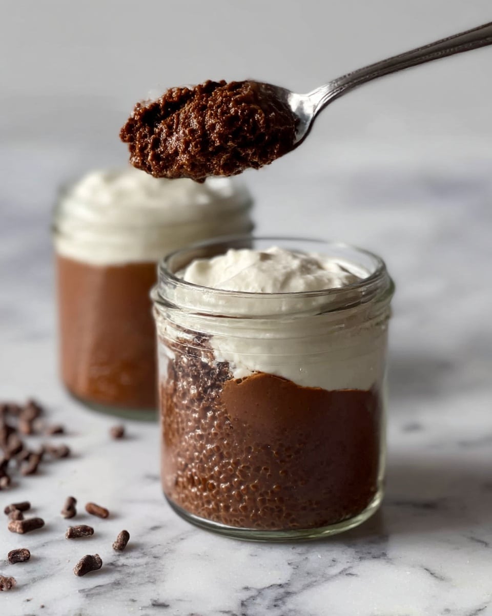 The image shows two small clear glass jars filled with a two-layer dessert. The bottom layer is a thick, dark brown chocolate pudding with a rough, slightly bumpy texture. On top, there is a smooth white cream layer that contrasts with the chocolate base. One jar is open with a silver spoon holding a scoop of the chocolate pudding and cream mix above it, showing the dense texture. The jars are placed on a white marbled surface with some scattered cacao nibs nearby. Photo taken with an iphone --ar 4:5 --v 7