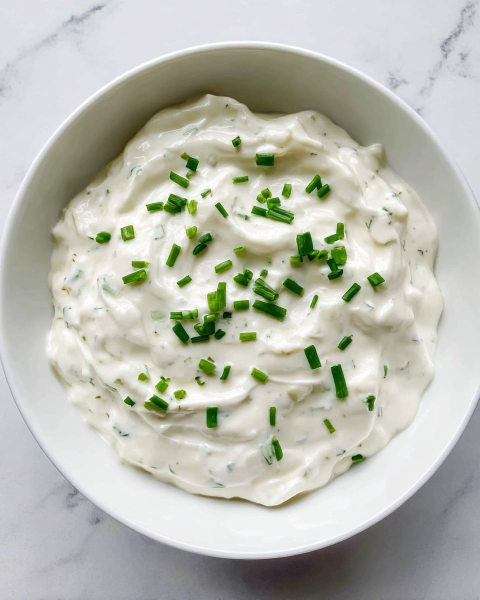 A white bowl filled with a creamy white dip that has a smooth and slightly thick texture. There are small green pieces of chopped chives sprinkled evenly on top, adding a pop of color and a fresh look. The bowl is placed on a white marbled surface, giving a clean and simple background. The light reflects softly on the surface of the dip, showing its smoothness and mild swirls. photo taken with an iphone --ar 4:5 --v 7