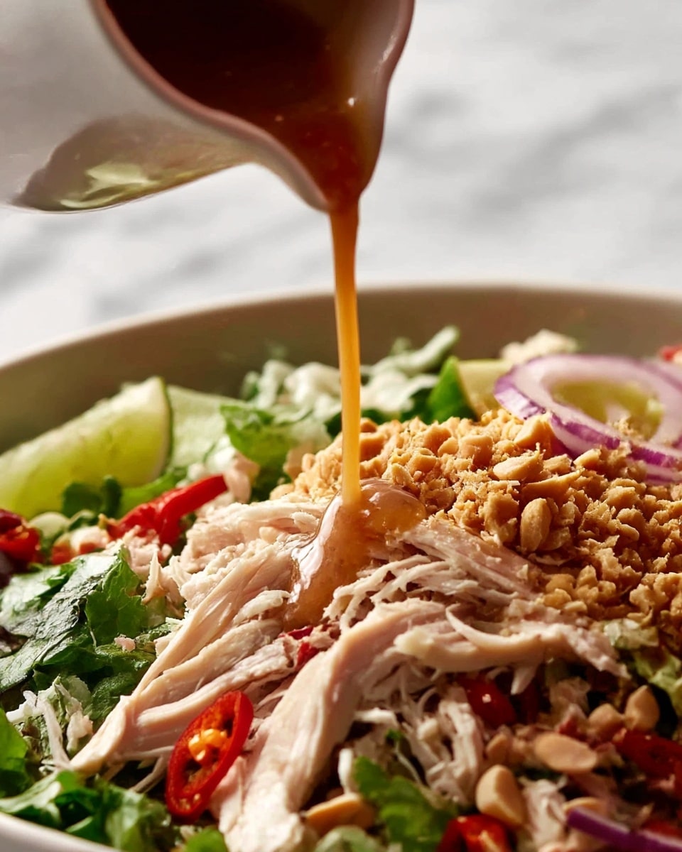 The image shows a close-up of a mixed salad that has several layers, starting with shredded white meat chicken on the bottom, mixed with green leafy herbs and thin red chili slices scattered throughout. On one side, there are crispy chopped peanuts creating a textured golden layer. Thinly sliced purple onions and bright lime wedges sit towards the back, adding color contrast. A light brown dressing is being poured from a white pitcher onto the salad, adding a shiny, smooth layer over the chicken and herbs. The background is a white marbled texture. photo taken with an iphone --ar 4:5 --v 7