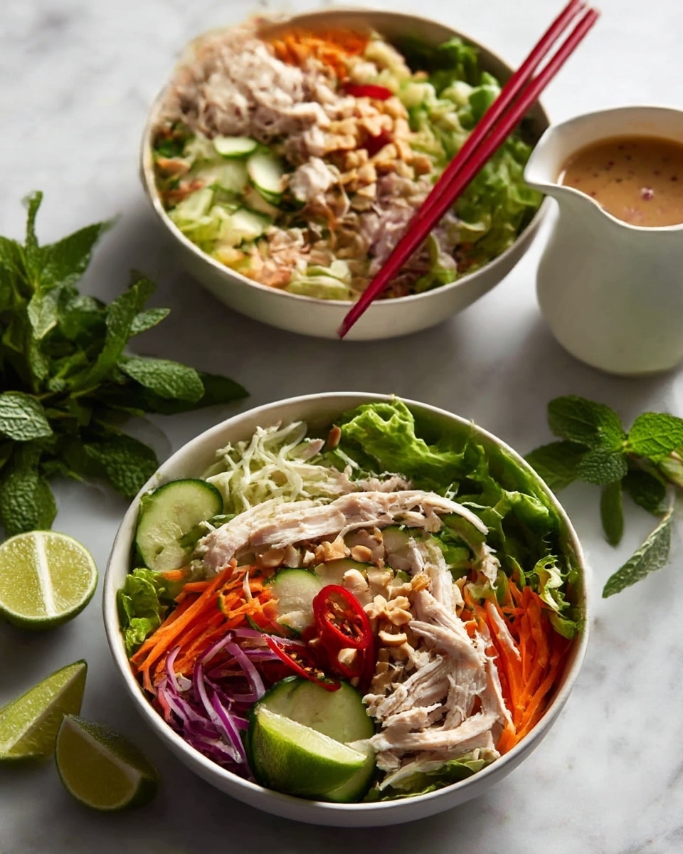 A large metal bowl filled with a fresh salad showing multiple layers starting with a base of green leafy lettuce, followed by thin slices of red bell pepper and shredded white cabbage. On top, there are shredded light brown chicken pieces, thin purple onion slices, and cucumber slices mixed in. The salad is garnished with chopped nuts, sprigs of fresh green mint and cilantro, and lime wedges placed on the side. A pair of wooden salad spoons rests inside the bowl. In the background on a white marbled surface, more fresh mint and a small white gravy boat filled with brown dressing can be seen. Photo taken with an iphone --ar 4:5 --v 7