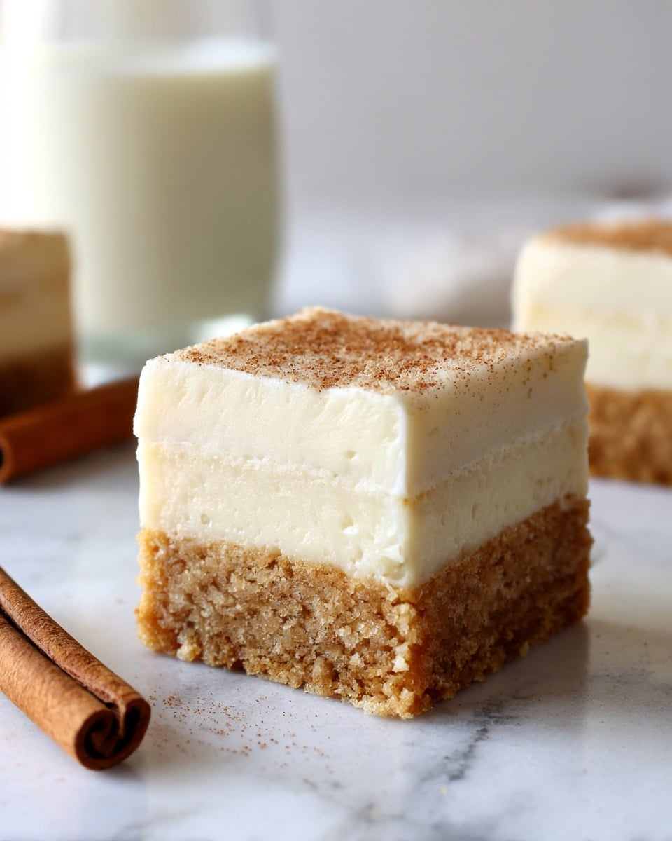 A close-up of a square dessert bar with two clear layers, the bottom layer is thick, light brown with a soft, crumbly texture, and the top layer is a thick, smooth, creamy white frosting evenly spread on top. The background shows a white marbled surface with a cinnamon stick and a glass of milk blurred in the back. Photo taken with an iphone --ar 4:5 --v 7