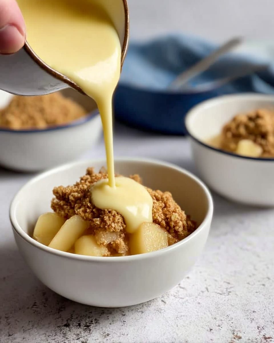 An oval-shaped dark baking dish filled with a single thick layer of golden-brown crumb topping that looks crisp and textured, showing small chunks and oats, covering the entire surface. The dish is placed on a light wooden board, which rests on a white marbled surface. Nearby, there are two empty white bowls stacked, and part of a blue, white, and grey checkered cloth is visible to the side. The crumb layer has an even, crunchy appearance with a warm toasted color. Photo taken with an iphone --ar 4:5 --v 7