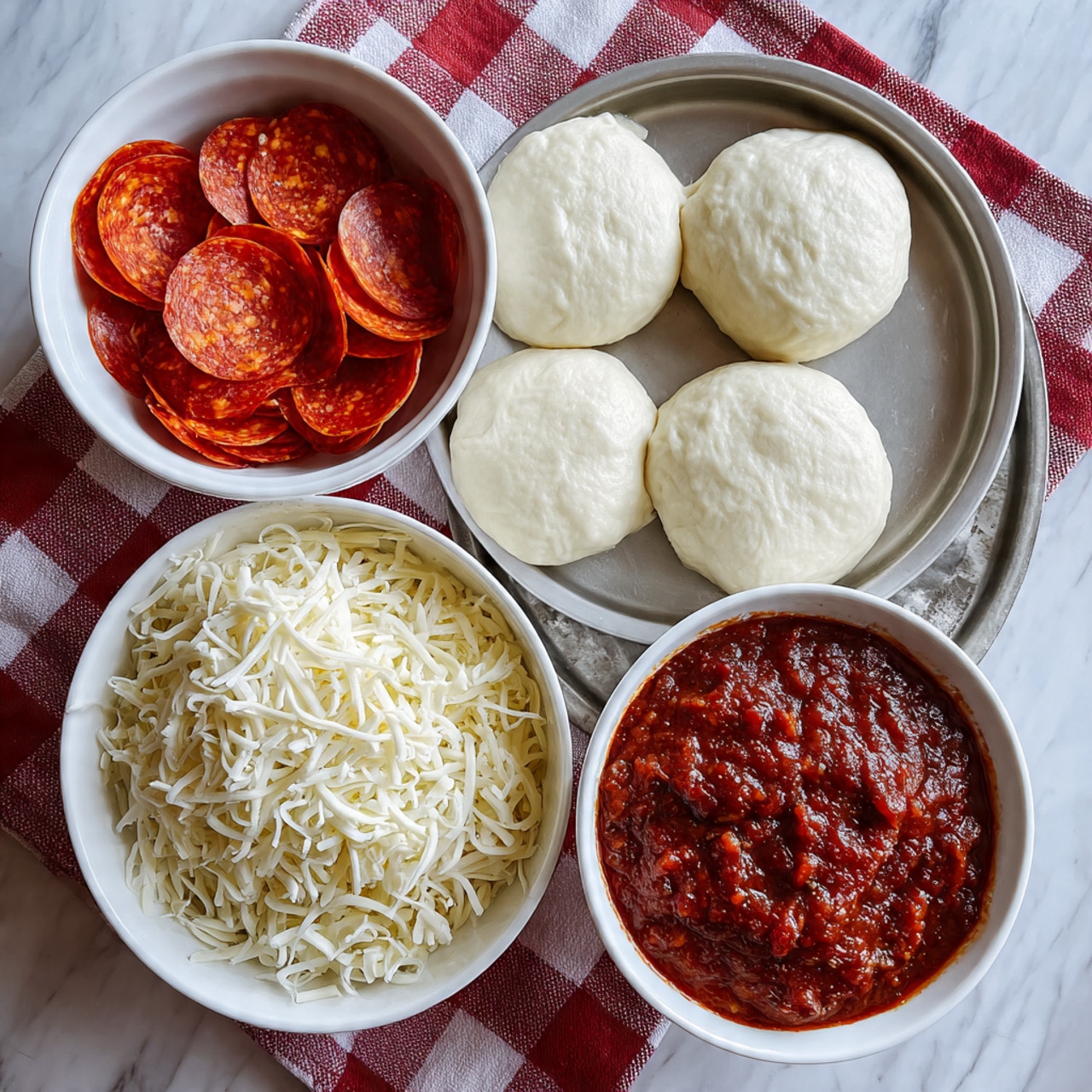 The image shows a metal tray with three layers of ingredients for making mini pizzas on a white marbled surface. On the top right of the tray, there are about eight round white dough pieces stacked in two layers, smooth and thick. Below the dough, covering the lower half of the tray, is a generous pile of shredded white cheese with a soft, slightly stringy texture. To the top left of the tray, there is a white bowl filled with bright red pepperoni slices stacked loosely, shiny with a slight oily surface. Next to it, a larger white bowl holds a rich, chunky red tomato sauce with visible bits of herbs and ingredients. Both bowls are placed on a red and white checkered cloth. photo taken with an iphone --ar 4:5 --v 7