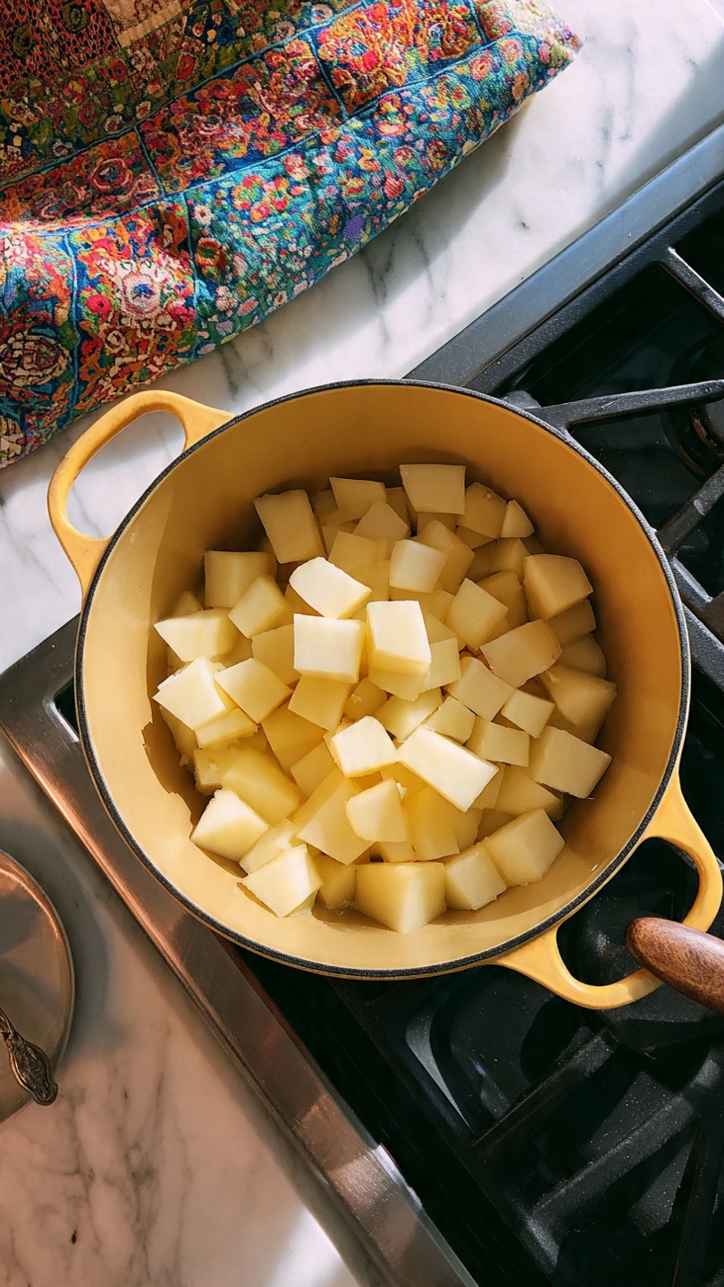 A yellow pot filled with several pieces of peeled potatoes, cut into small cubes, sitting over a black stove with visible burner grates, all placed on a white marbled surface with part of a colorful cloth seen to the left side. photo taken with an iphone --ar 4:5 --v 7