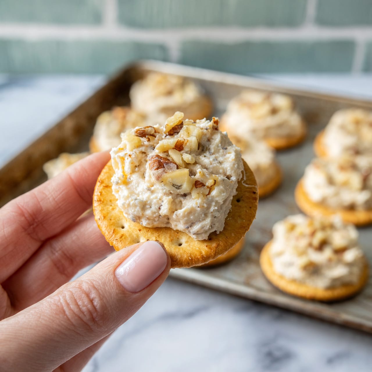 A close-up image shows a woman's hand holding a round golden cracker topped with a creamy beige mixture that has small nut pieces mixed in. In the background, a metal baking sheet holds several more crackers with the same topping, arranged in rows. The surface underneath is a white marbled texture, and the background is made of light green painted bricks. The lighting is soft, highlighting the texture of the cracker and creamy topping. photo taken with an iphone --ar 4:5 --v 7