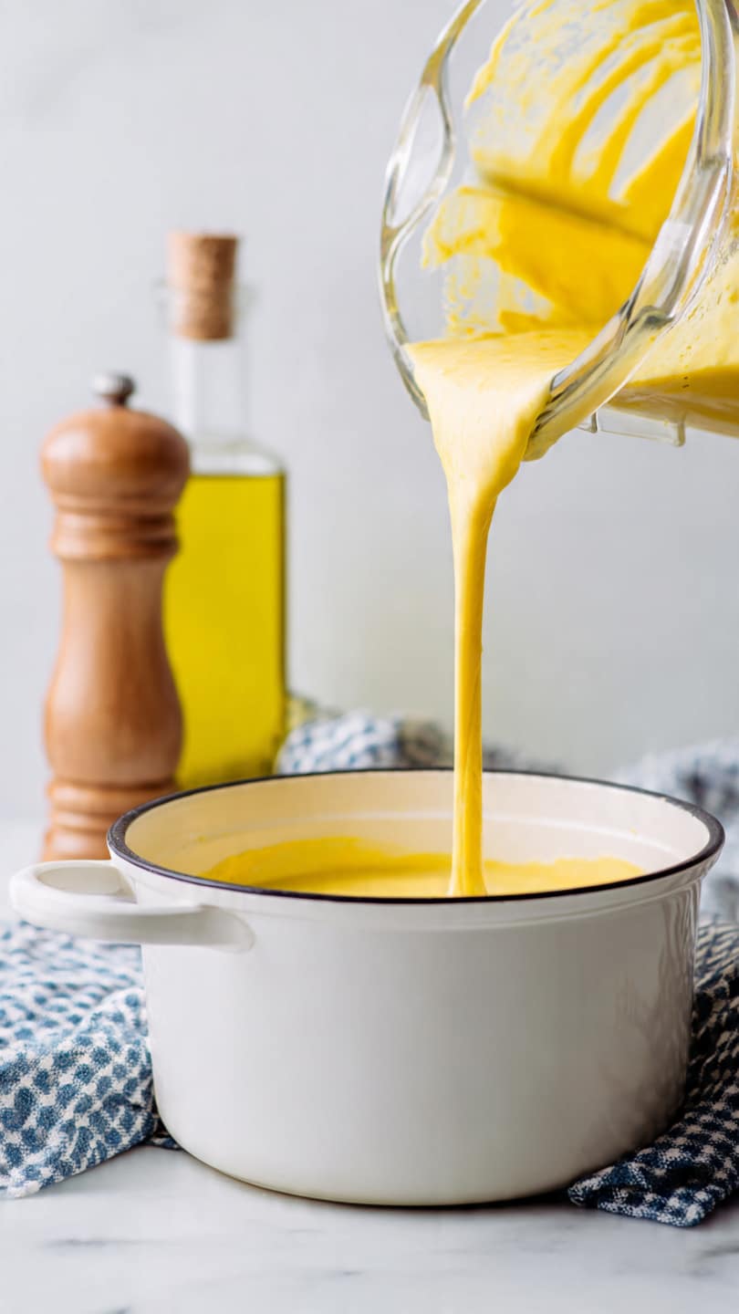 A close-up view shows thick smooth yellow batter being poured from a clear blender container into a white pot with black inside, placed on a white marbled surface. In the background, there is a tall bottle of yellow oil and a light wooden pepper grinder next to a folded blue and white checkered cloth. Steam rises from the pot, hinting the contents inside are hot. The scene is bright and clean with soft lighting highlighting the pouring motion. Photo taken with an iphone --ar 4:5 --v 7