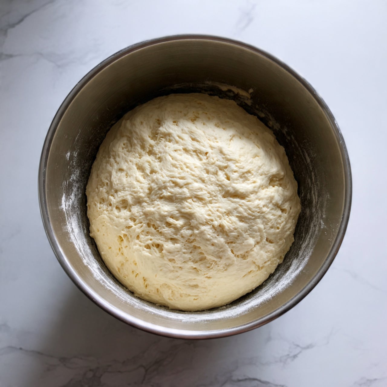 A soft, round bun is sliced in half horizontally with the top half dusted with a light layer of white powdered sugar. Between the two halves is a thick, smooth layer of white cream that swirls outward slightly beyond the bun edges, showing a light and fluffy texture. The bun sits centered on a white plate decorated with pink and green floral patterns, all placed on a surface with a white marbled texture. photo taken with an iphone --ar 4:5 --v 7