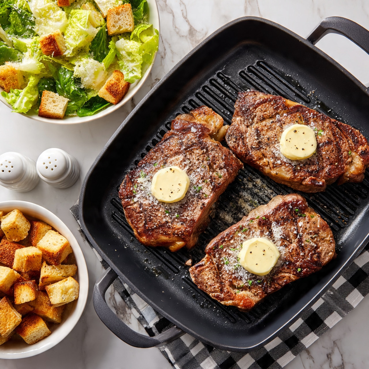 The image shows three cooked steaks placed side by side on a rectangular white plate. Each steak has a small round scoop of butter melting on top, with visible pepper seasoning on the brown, slightly charred surface. The plate is set on a white marbled wooden table, with two white salt and pepper shakers near the top and green parsley sprigs on the sides. To the left, there is a white bowl filled with a green salad that includes lettuce, croutons, and shaved cheese. A black and white checkered cloth is partially visible under the bowl. On the right edge, a white plate with two forks rests on the table. Photo taken with an iphone --ar 4:5 --v 7