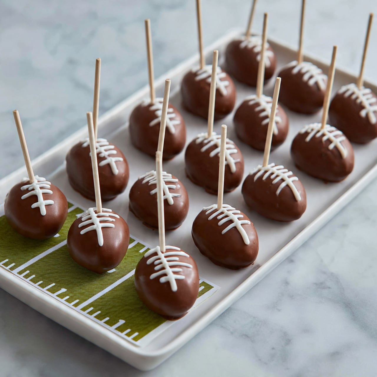 The image shows a white rectangular plate on a white marbled surface holding twelve small football-shaped treats. Each treat is covered in smooth milk chocolate forming a deep brown, shiny shell. On top of each, there are four short white icing lines crossing a longer white horizontal icing line, mimicking football laces. Each treat has a white stick inserted at the narrow end, standing upright. The treats are arranged in rows, neatly filling the plate. Part of a green paper football field with white markings can be seen under the plate. photo taken with an iphone --ar 4:5 --v 7