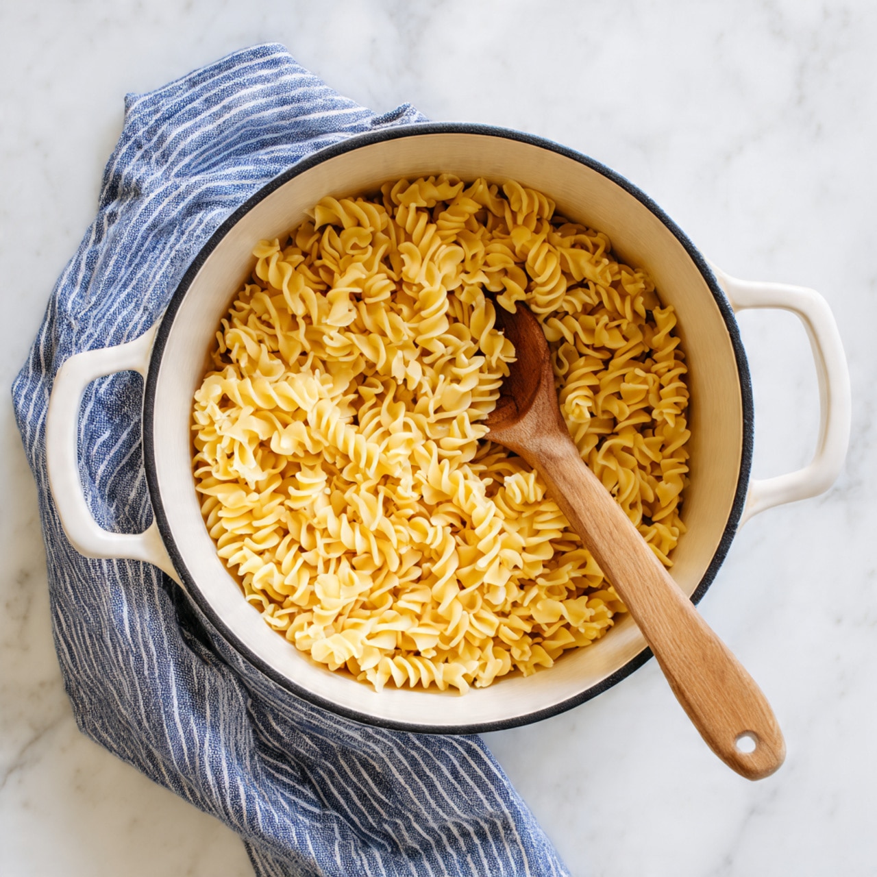 A white pot filled with uncooked twisted egg noodles that are golden yellow in color, filling almost the entire pot. A wooden spoon with a long handle is inside the pot, resting on the noodles towards the center. The pot has a black rim and two handles on each side. The pot is placed on a white marbled surface, next to a blue and white striped kitchen towel laid casually to the left side. photo taken with an iphone --ar 4:5 --v 7