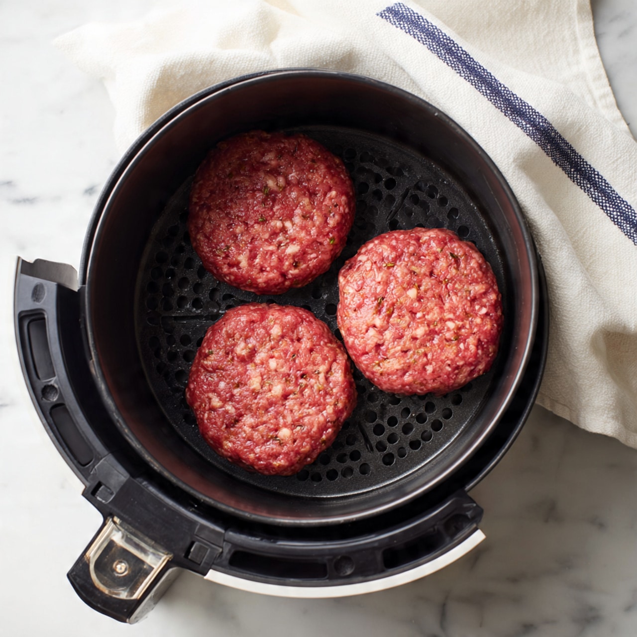 Inside a black round air fryer basket with small holes along its sides, three raw hamburger patties sit spaced evenly. Each patty has a reddish-pink color with visible small white fat bits and rough textured surface. The air fryer basket is set on a white marbled surface with a white cloth towel with blue stripes next to it. The air fryer basket's handle is visible at the bottom of the image. photo taken with an iphone --ar 4:5 --v 7