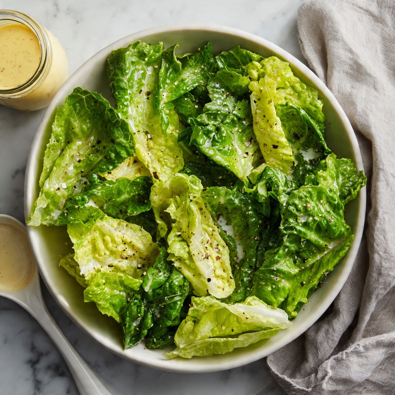 A white bowl filled with bright green leafy lettuce that looks fresh and slightly shiny with dressing drizzled over it. Small bits of black pepper and light dressing dots are scattered on the lettuce leaves. Next to the bowl is a small glass jar of light creamy yellow dressing and a white round lid with a spoon resting on it. The whole scene is set on a white marbled surface with a crumpled light grey cloth on the right side of the image. photo taken with an iphone --ar 4:5 --v 7