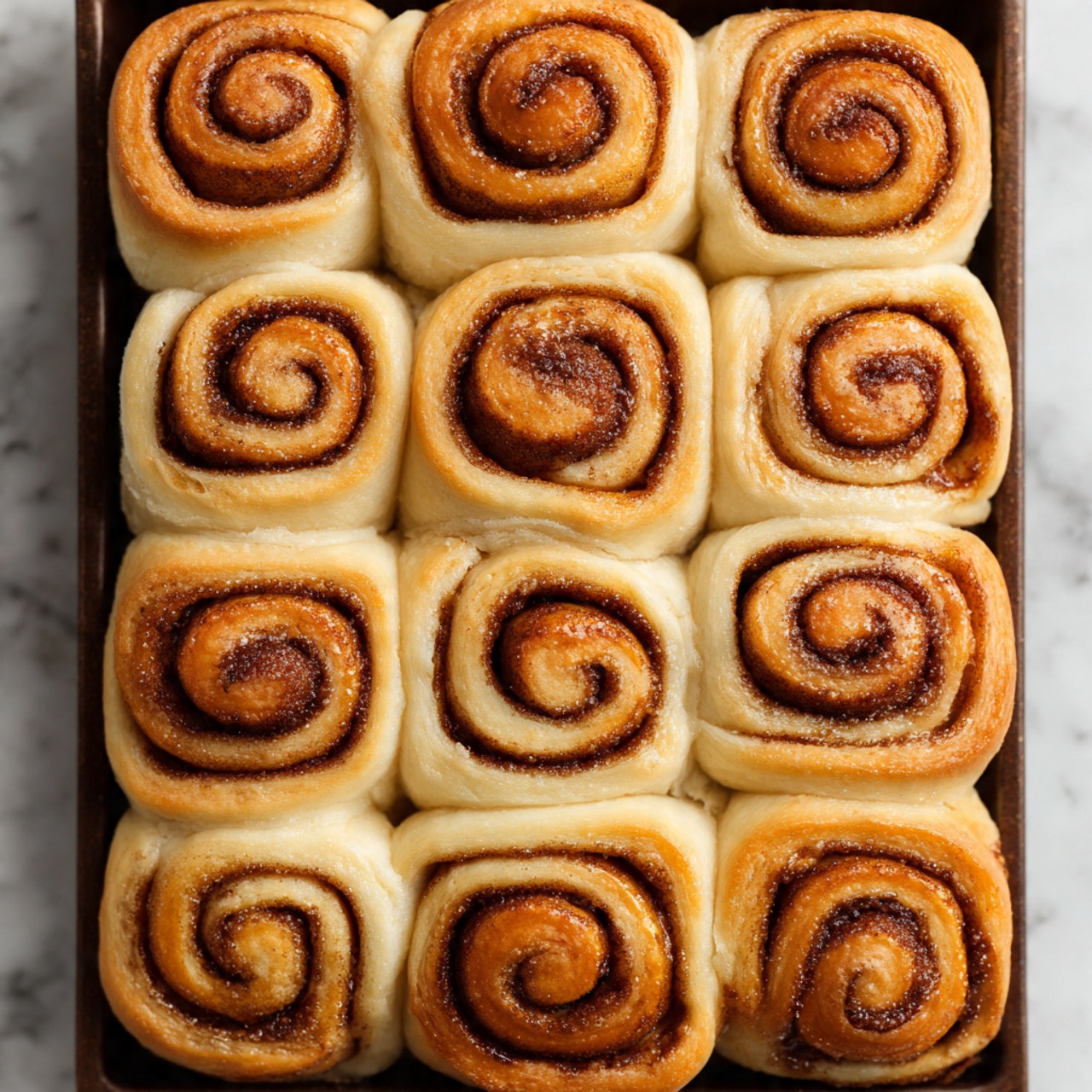 A close-up top view of a tray filled with 15 cinnamon rolls arranged in a 3 by 5 grid. Each roll shows a distinct spiral pattern with a golden brown top layer that has a slightly glossy texture, highlighting the cinnamon swirl inside. The rolls are soft and fluffy with a light beige dough base visible around the cinnamon spiral. They are snugly placed next to each other in a dark baking tray, which contrasts with the light color of the rolls. The background is a white marbled texture. photo taken with an iphone --ar 4:5 --v 7
