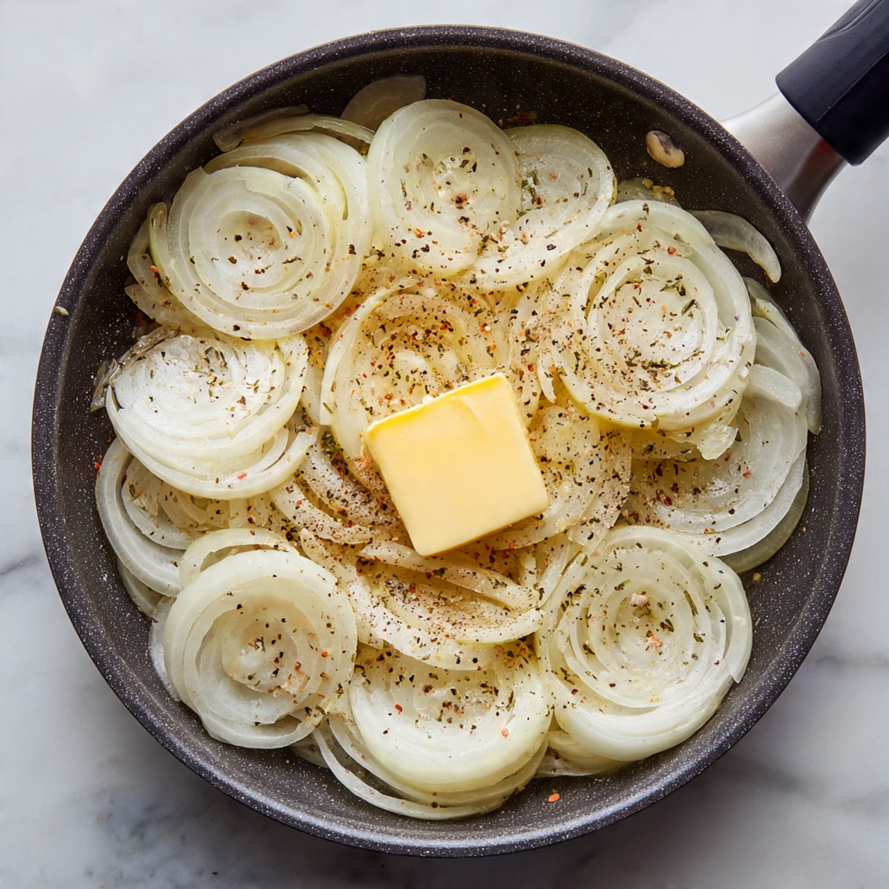 A dark non-stick pan filled with a single layer of pale sliced onions that show their curved rings clearly, topped in the center with a square piece of yellow butter melting slowly. The onions are sprinkled evenly with coarse black pepper and some light pink salt granules. The pan sits on a white marbled surface, showing the simple ingredients ready to be cooked. photo taken with an iphone --ar 4:5 --v 7