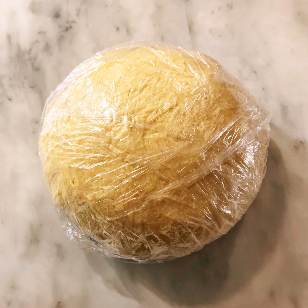 This image shows a slice of pie on a white plate placed on a folded dark gray cloth over a white marbled surface. The pie has three layers: a light golden crust on the bottom with a crumbly texture, a thick layer of smooth caramel brown filling in the middle, and a thick layer of fluffy white whipped cream on top. Small toffee or nut bits are sprinkled on the whipped cream. A spoon with some pie on it rests on the right side of the plate. In the background, parts of the whole pie and another slice are blurred but show the same layers. Photo taken with an iphone --ar 4:5 --v 7