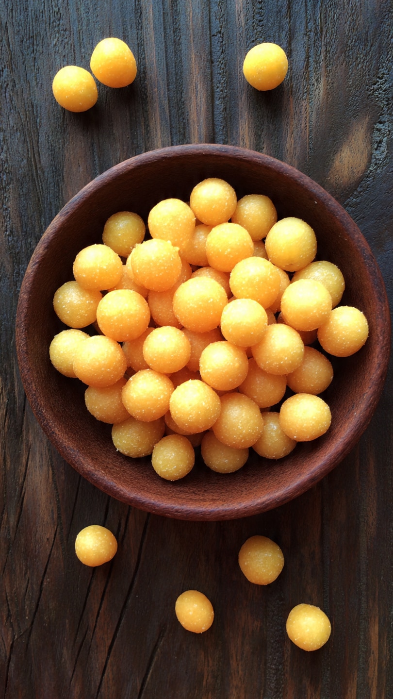A brown bowl filled with small, round, yellow puffed balls arranged close together inside, with several loose pieces scattered around it on a dark wooden surface. The balls have a smooth and slightly shiny texture, looking light and crunchy. photo taken with an iphone --ar 4:5 --v 7