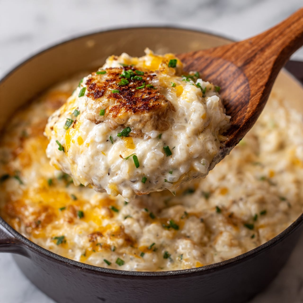A close-up view of a creamy yellow cheesy rice dish with visible soft cooked rice grains mixed with melted cheese, and tender browned chicken pieces scattered throughout. The dish is inside a blue cooking pot with a wooden spoon stirring it from the left side. The texture is smooth and creamy, with some black pepper specks on the cheese. The background is a white marbled surface. Photo taken with an iphone --ar 4:5 --v 7