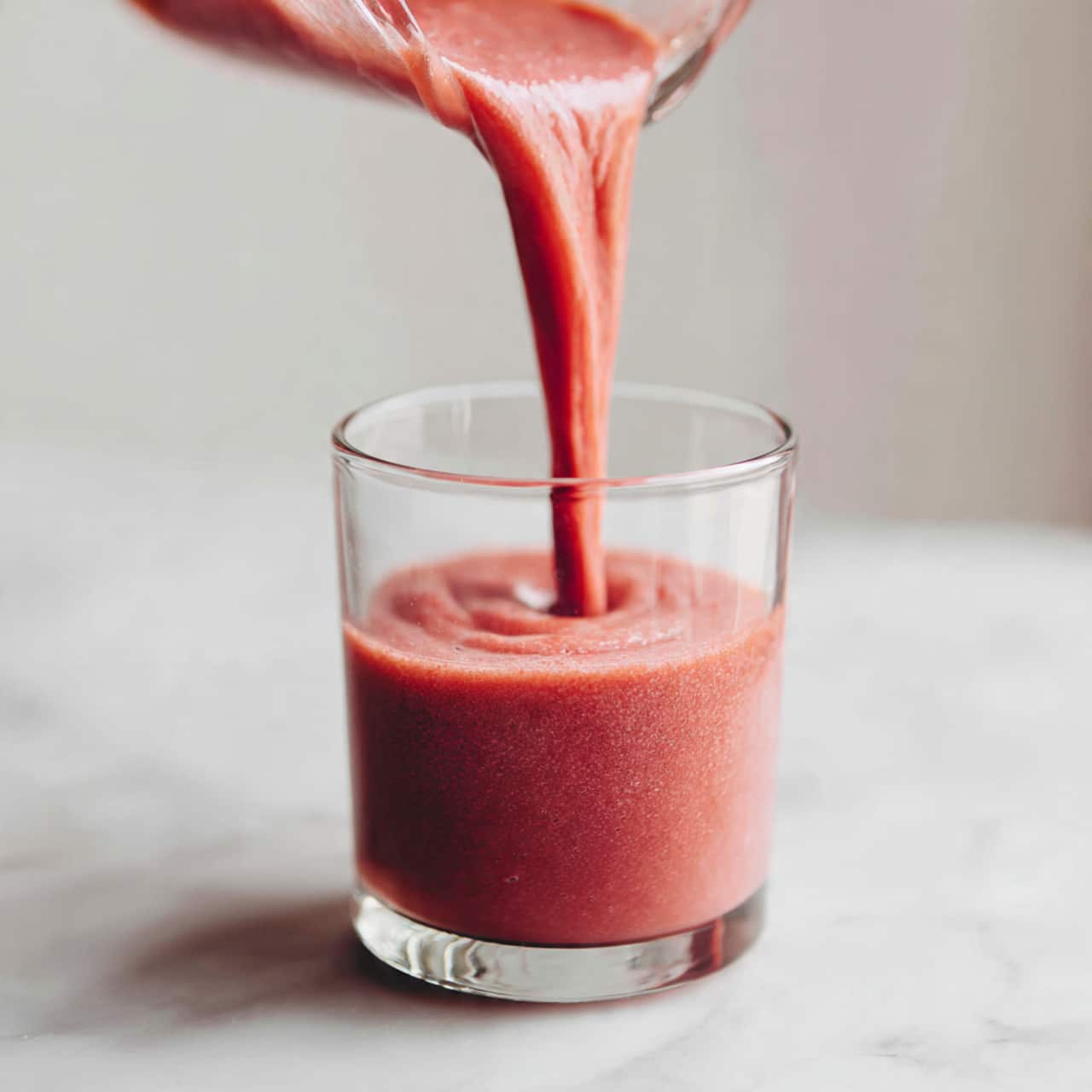 A clear glass cup filled with two thick layers: the bottom layer is a rich, red velvet mousse with a soft, slightly textured surface, and the top layer is a fluffy white whipped cream that peaks high above the rim, dusted with small crumbs of red velvet cake. The cup sits on a white marbled surface next to another similar cup. The background is softly blurred with a light, neutral tone. Photo taken with an iphone --ar 4:5 --v 7