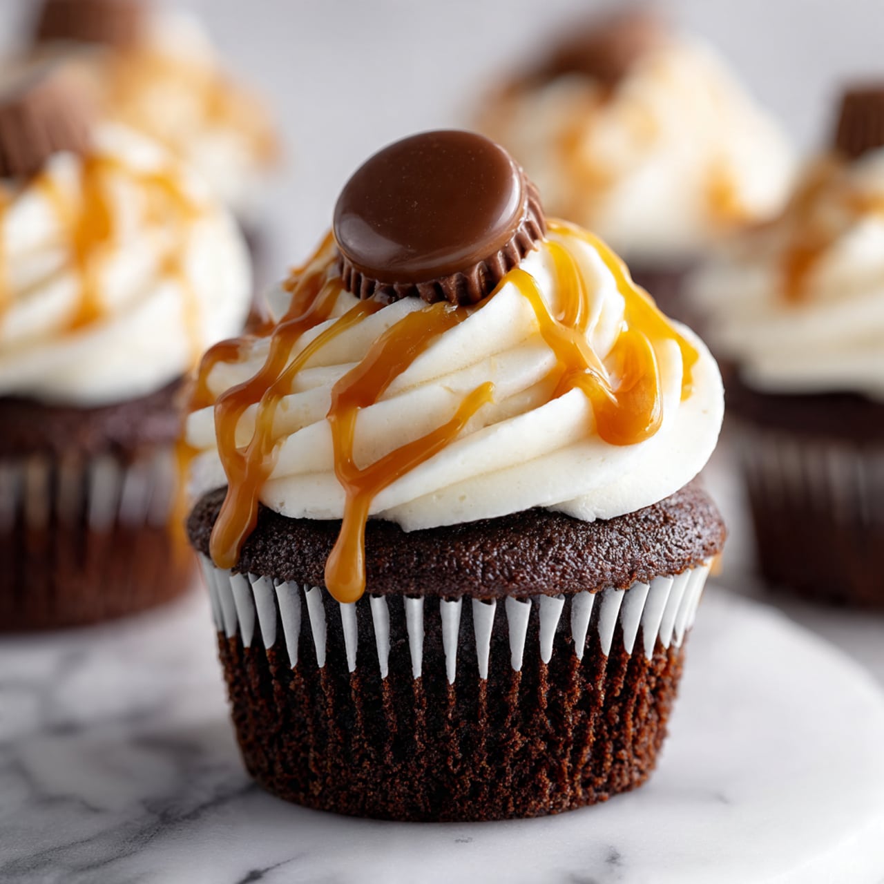 A single chocolate cupcake with a dark brown moist base sits on a white marbled surface. On top, there is a thick layer of smooth white frosting swirled in soft peaks, generously drizzled with golden caramel sauce. A half chocolate-covered caramel candy with a creamy inside is placed on the very top, centered on the frosting. The background shows blurred similar cupcakes with white frosting and caramel drizzle. Photo taken with an iphone --ar 4:5 --v 7