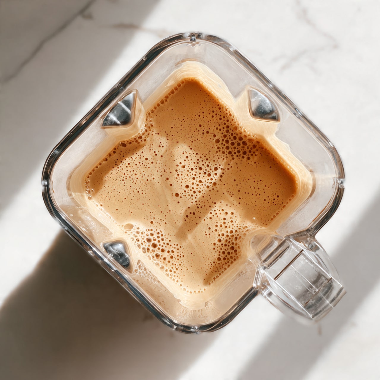 A clear blender container filled with a light tan thick liquid that has small bubbles on its smooth surface, seen from above. The blender sits on a white marbled surface, with light casting soft shadows to the side. The container has a handle on the right side and distinct angled shapes inside the blender. The mixture appears creamy and well-blended, with no visible chunks or pieces. photo taken with an iphone --ar 4:5 --v 7