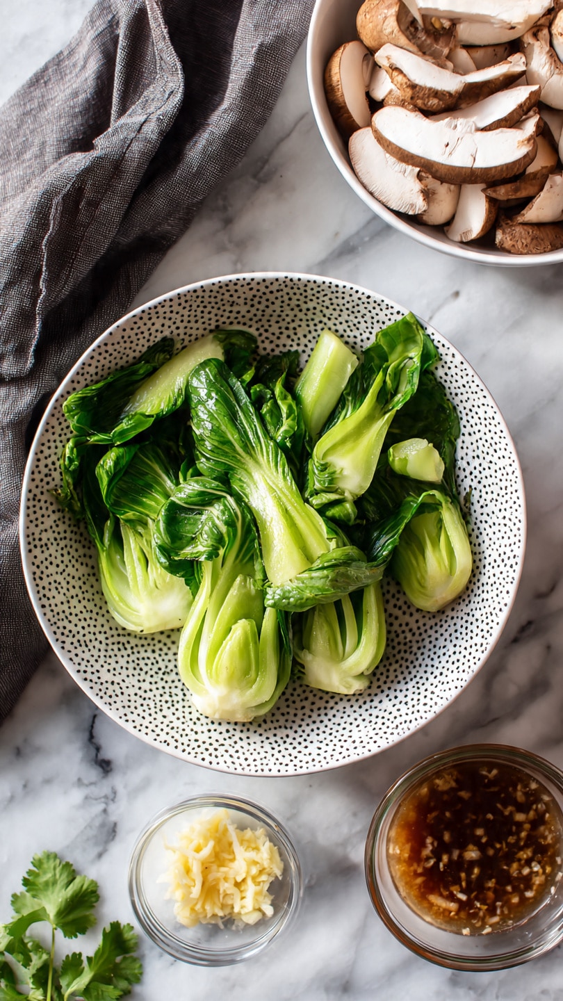 The image shows a white patterned bowl filled with fresh green bok choy, some whole and some cut into pieces, positioned in the center on a white marbled surface. Above and slightly to the left is a white bowl containing light brown sliced mushrooms, layered neatly. Below and to the right of the bok choy bowl is a small clear glass dish with finely grated yellow ginger next to a glass container filled with a brown sauce with visible bits. A small sprig of cilantro rests on the surface near the bottom left corner. A dark gray cloth is draped softly on the left side. photo taken with an iphone --ar 4:5 --v 7