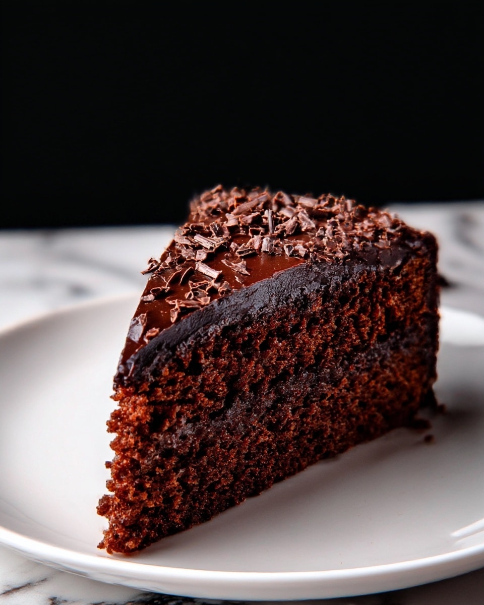 A dark chocolate cake sits on a white plate, showing one thick layer fully covered with a shiny, smooth chocolate glaze. The top is sprinkled with small, rough chocolate shavings spread evenly. The cake has a rich, moist texture with a slightly bumpy side surface under the glossy glaze. The background is plain black which contrasts with the white plate and the dark cake. photo taken with an iphone --ar 4:5 --v 7