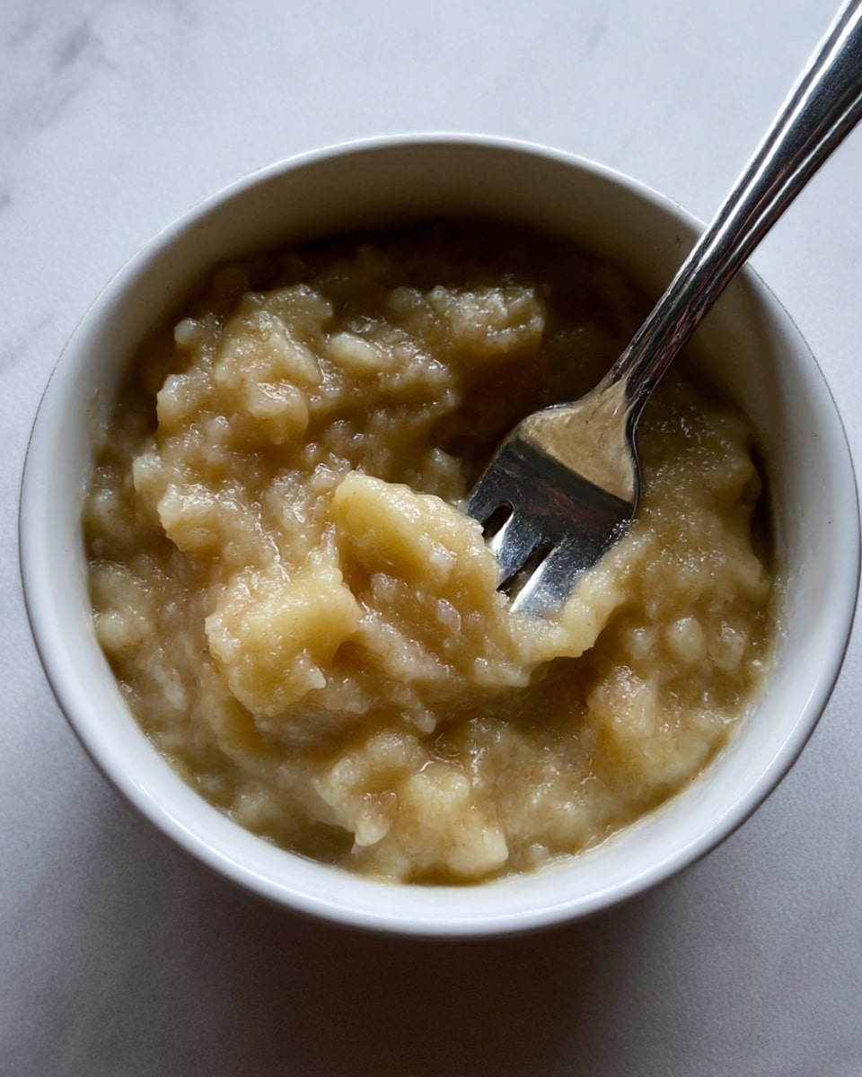 A close-up view of a small white bowl filled with mashed bananas that have a soft, lumpy texture and a pale yellow-brown color. A metal fork with a shiny silver handle is resting inside the bowl, slightly mixed into the mashed bananas. The background surface is a clean white marbled texture. photo taken with an iphone --ar 4:5 --v 7