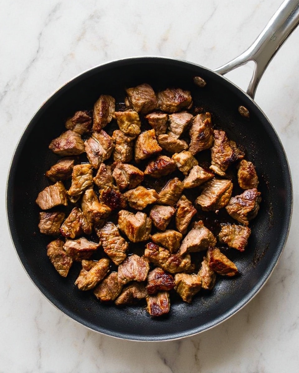 The image shows a black frying pan filled with several pieces of cooked meat. The meat pieces are uneven in size and shape, showing a mix of brown, golden, and slightly charred colors, indicating they are seared and cooked. The pan has a silver handle, and the background is a white marbled surface. The meat pieces are spread so they cover the pan but do not overlap much, creating a textured and hearty look. photo taken with an iphone --ar 4:5 --v 7