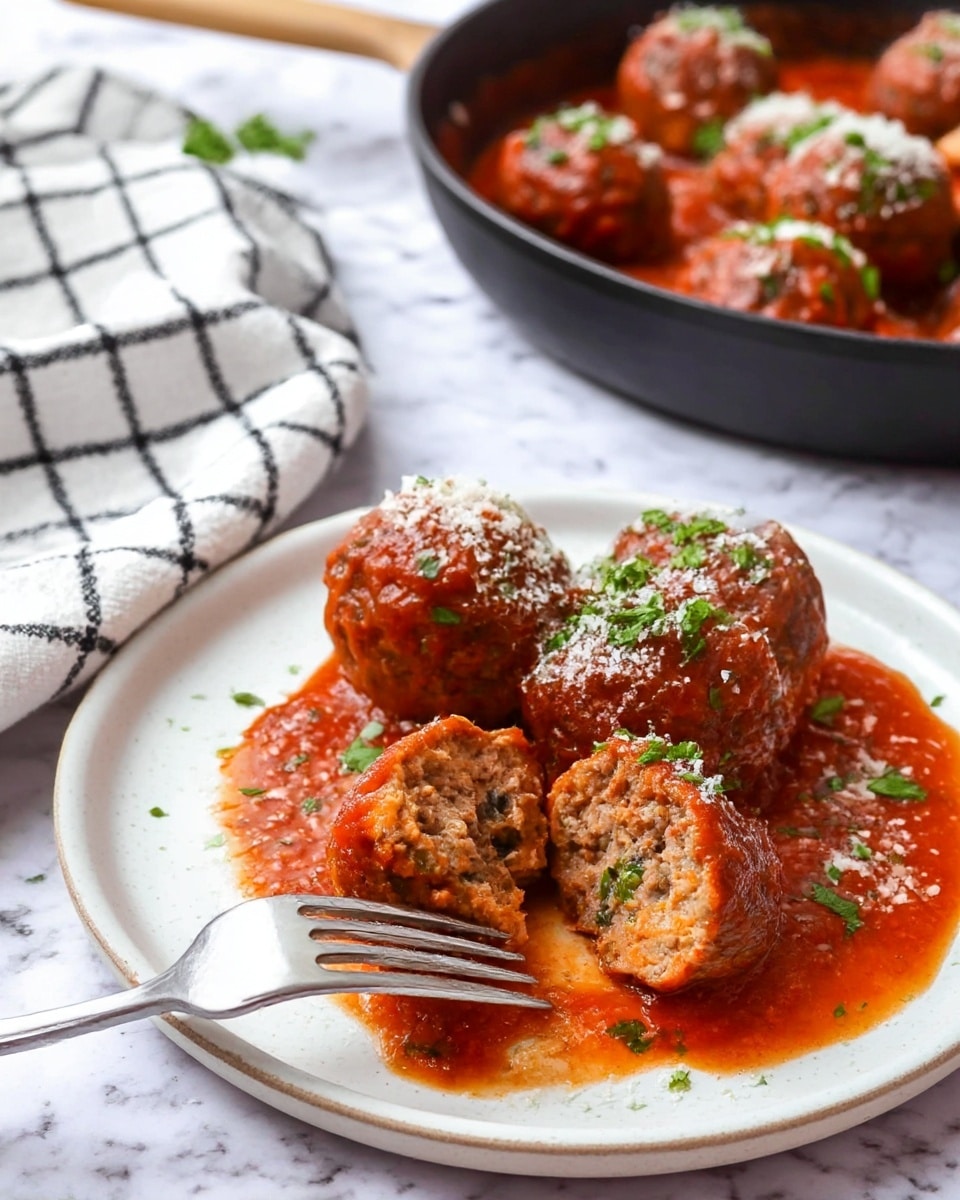 The image shows a white plate with four big round meatballs, each covered in a thick red tomato sauce. One meatball is cut open, showing its soft, cooked inside with small green herb bits. The meatballs are topped with green chopped herbs and a light sprinkle of white grated cheese. The tomato sauce pools around the meatballs with a slightly oily shine. A fork rests on the plate, holding the cut meatball. In the background, there is a black pan filled with more meatballs in sauce, sprinkled with herbs. The scene is set on a white marbled surface with a folded white and dark checkered cloth nearby. photo taken with an iphone --ar 4:5 --v 7