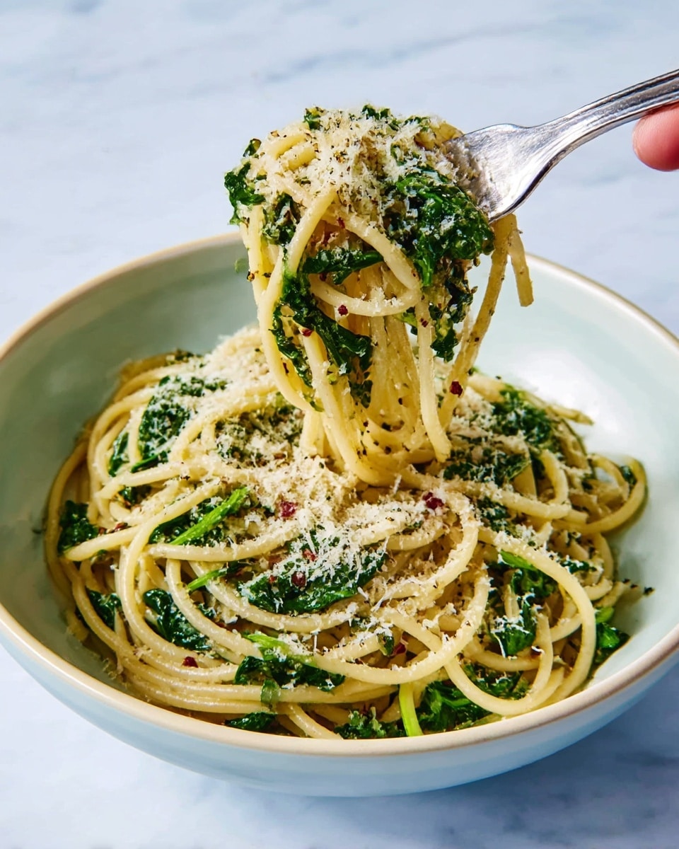 A white bowl holds a serving of spaghetti mixed with fresh green spinach leaves and small bits of red chili flakes. The pasta is coated with a light sauce and sprinkled generously with finely grated pale yellow cheese and black pepper, showing specks all over. A silver fork held by a woman's hand lifts some spaghetti strands mixed with spinach and cheese above the bowl. The background is a white marbled surface. photo taken with an iphone --ar 4:5 --v 7