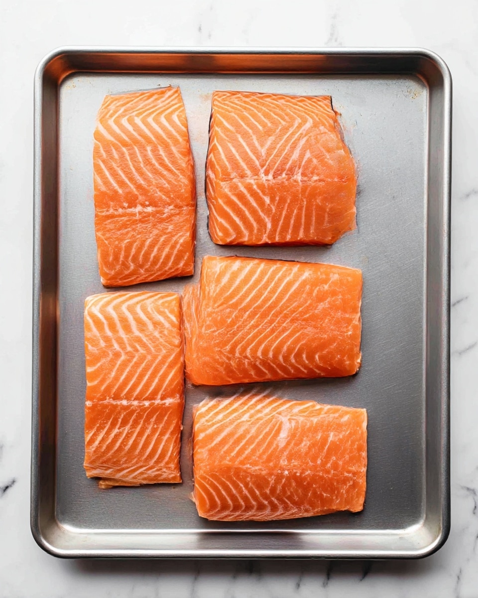 The image shows a metal baking tray placed on a white marbled surface, holding four raw salmon fillets arranged in a neat vertical row. Each fillet has a rich orange color with visible white fat lines running through them, creating a striped pattern. The texture of the salmon is smooth with slight wetness, and the fillets are similar in size and shape, lying flat on the tray with no additional ingredients. The overall scene is clean and simple, focused on the fresh salmon ready for cooking photo taken with an iphone --ar 4:5 --v 7