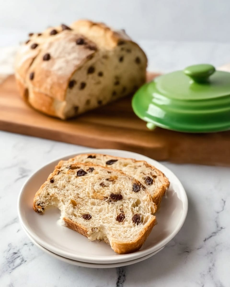 Two slices of bread with dark bits inside are placed on a white plate. The front slice has a small bite taken out of it, showing its soft texture. Behind the plate, there is a round loaf of bread with the same dark bits, resting on a wooden board. Next to the loaf, there is a green butter dish with the lid on. The surface under the plate and board is white with a marbled texture. photo taken with an iphone --ar 4:5 --v 7
