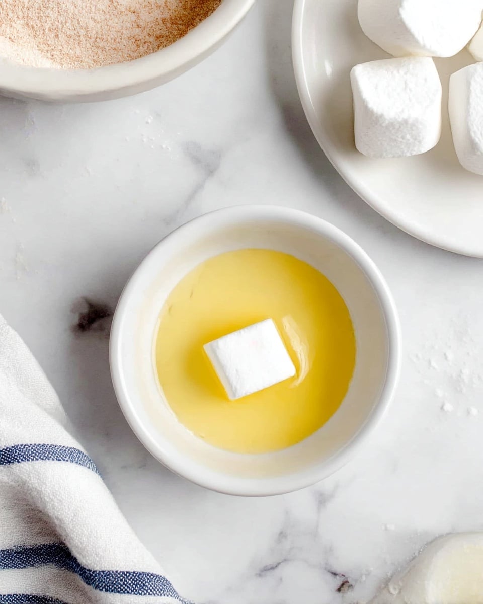 A small white bowl with a single white marshmallow sitting in a pool of melted yellow butter. Near it, on a white marbled surface, there is a white plate holding several other white marshmallows, and part of a white bowl with light brown powder inside is visible in the top left corner. A white and blue striped cloth is partially shown at the bottom left. Photo taken with an iphone --ar 4:5 --v 7