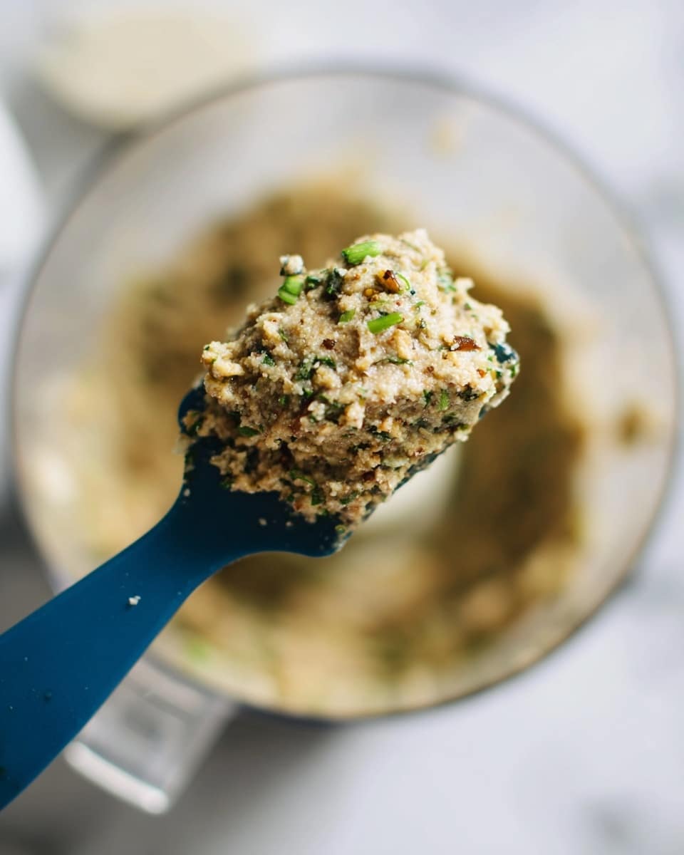 A close-up of a blue spatula holding a thick, coarse mixture with small bits of green herbs and darker brown pieces, showing a textured, uneven surface. In the background, a clear food processor bowl filled with the same mixture sits on a white marbled surface, slightly out of focus. The overall color of the mixture is light beige with green and brown specks scattered throughout. Photo taken with an iphone --ar 4:5 --v 7