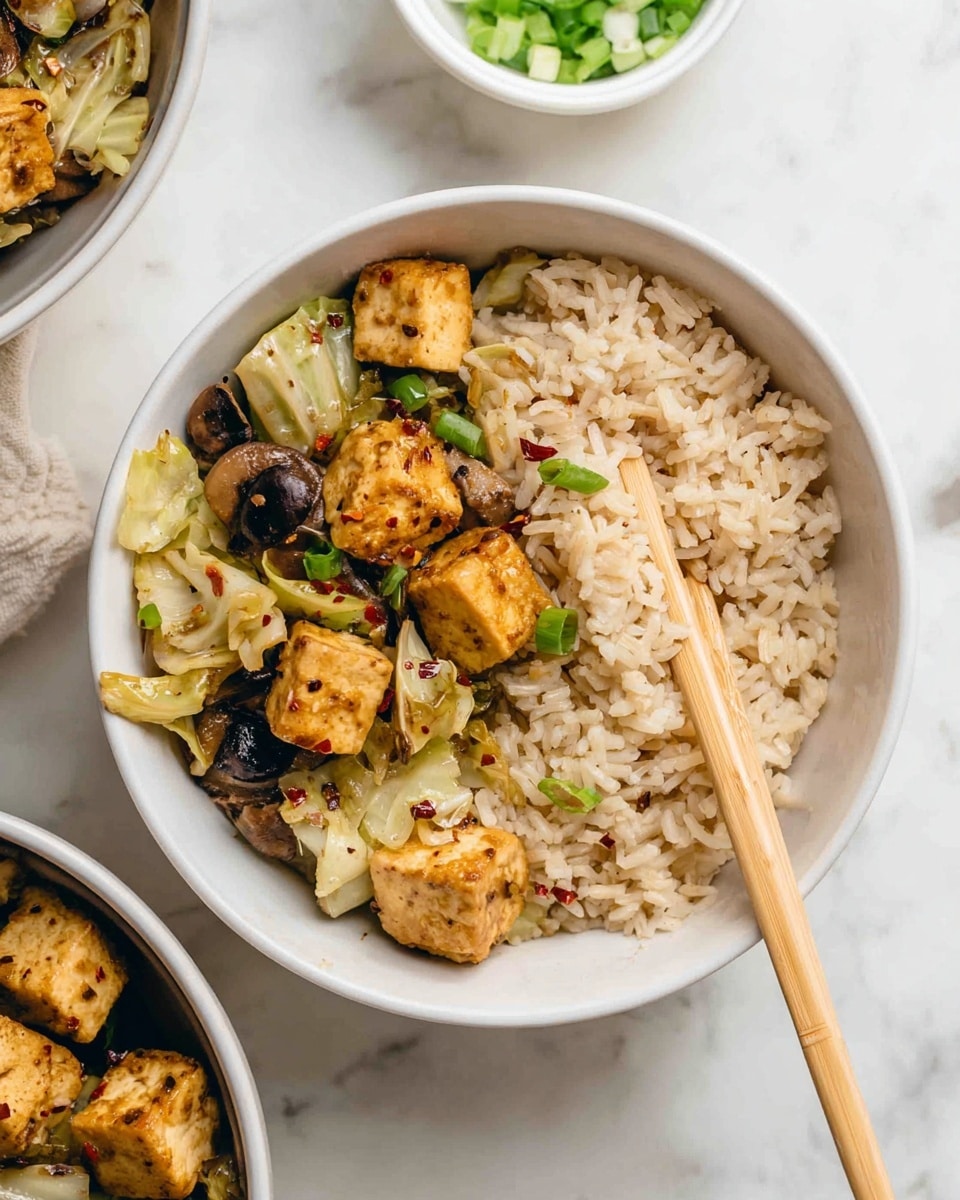 A white bowl filled with a stir-fry dish, showing about three main layers: golden brown tofu cubes on top, mixed with light brown, slightly curved mushroom slices, and pale green sliced cabbage throughout. The dish is sprinkled with small green onion pieces and red chili flakes, adding little pops of green and red colors. A woman's hand using light wooden chopsticks is picking up some pieces from the left side of the bowl. Around the bowl are small white dishes containing chopped green onions and red chili flakes, plus a separate bowl of cooked brown rice below on a white marbled surface. photo taken with an iphone --ar 4:5 --v 7