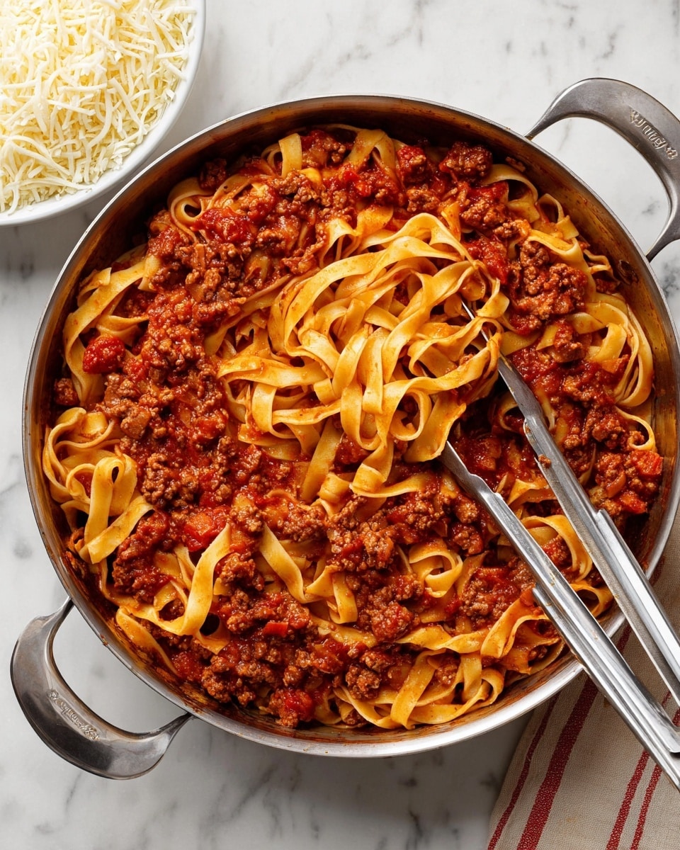 The image shows a large silver pan filled with fettuccine pasta mixed in a thick, rich red meat sauce. The sauce contains visible chunks of cooked ground meat spread evenly throughout the yellowish flat pasta ribbons, which have curled edges and show a slightly shiny texture from the sauce. A pair of silver tongs rests on the pasta near the bottom center of the pan. To the top right of the pan, there is a white bowl filled with shredded white cheese, all set on a white marbled surface. The overall look is warm and hearty, with a focus on the mixed pasta and meat sauce. photo taken with an iphone --ar 4:5 --v 7