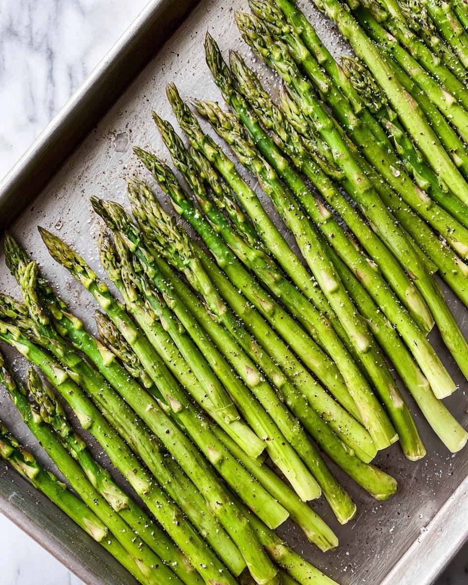 The image shows a single metal baking sheet filled with one layer of fresh, green asparagus spears. The asparagus is arranged in neat, nearly parallel rows covering the whole surface of the tray. The spears have light green stalks with darker green tips that are tightly closed and slightly textured. There are small black pepper flakes and a bit of coarse salt sprinkled evenly over the asparagus. The baking sheet has a slightly worn, silver metallic surface with some light scratches and a matte finish. The background is a white marbled texture. Photo taken with an iphone --ar 4:5 --v 7