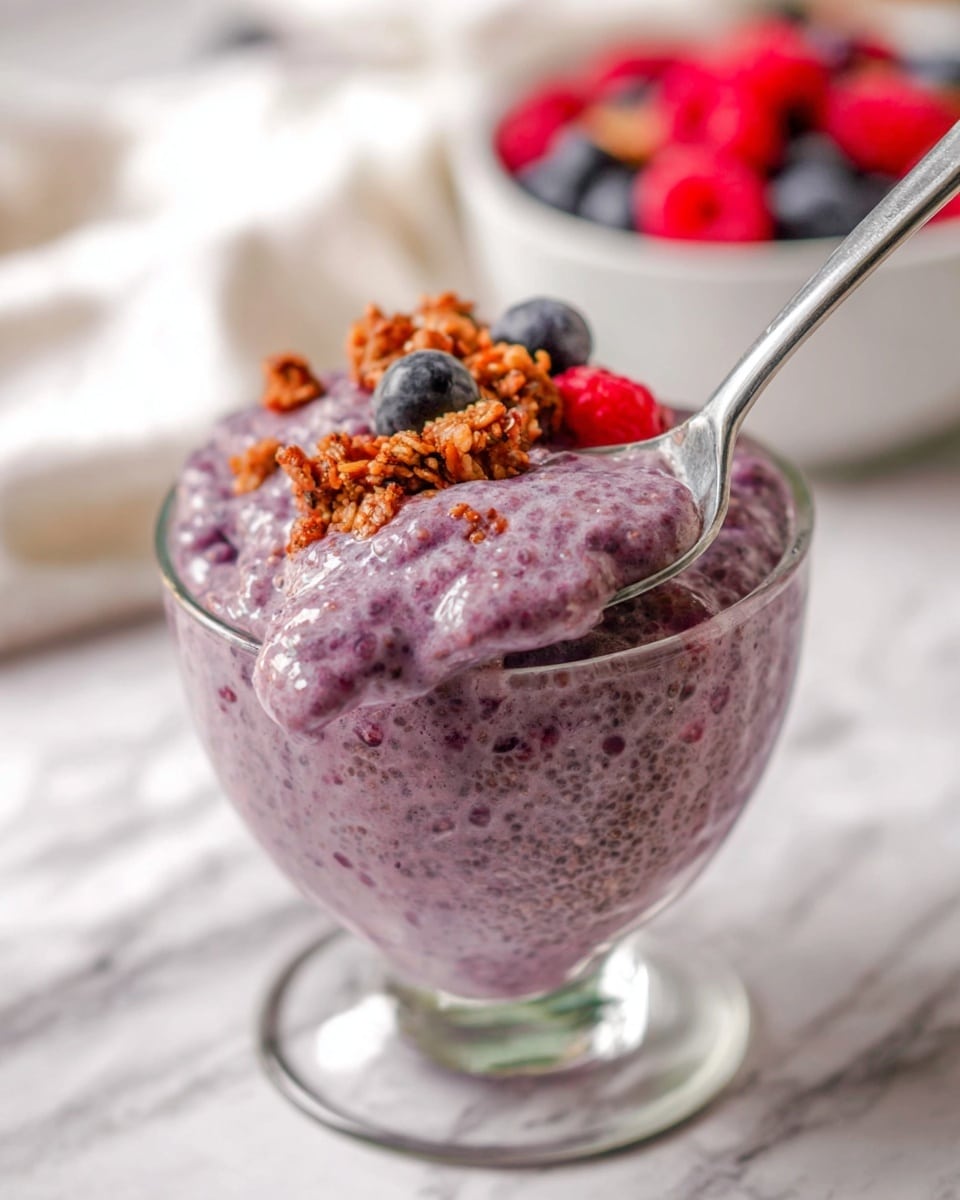 A round clear glass bowl filled with a smooth, thick pink smoothie that fills the bowl nearly to the top. The surface of the smoothie is speckled with small air bubbles. On top, near the center, there is a loose cluster of small chia seeds in a slightly irregular oval shape, dark brown and greenish in color, adding texture contrast. The bowl sits on a white marbled surface, seen from above. photo taken with an iphone --ar 4:5 --v 7