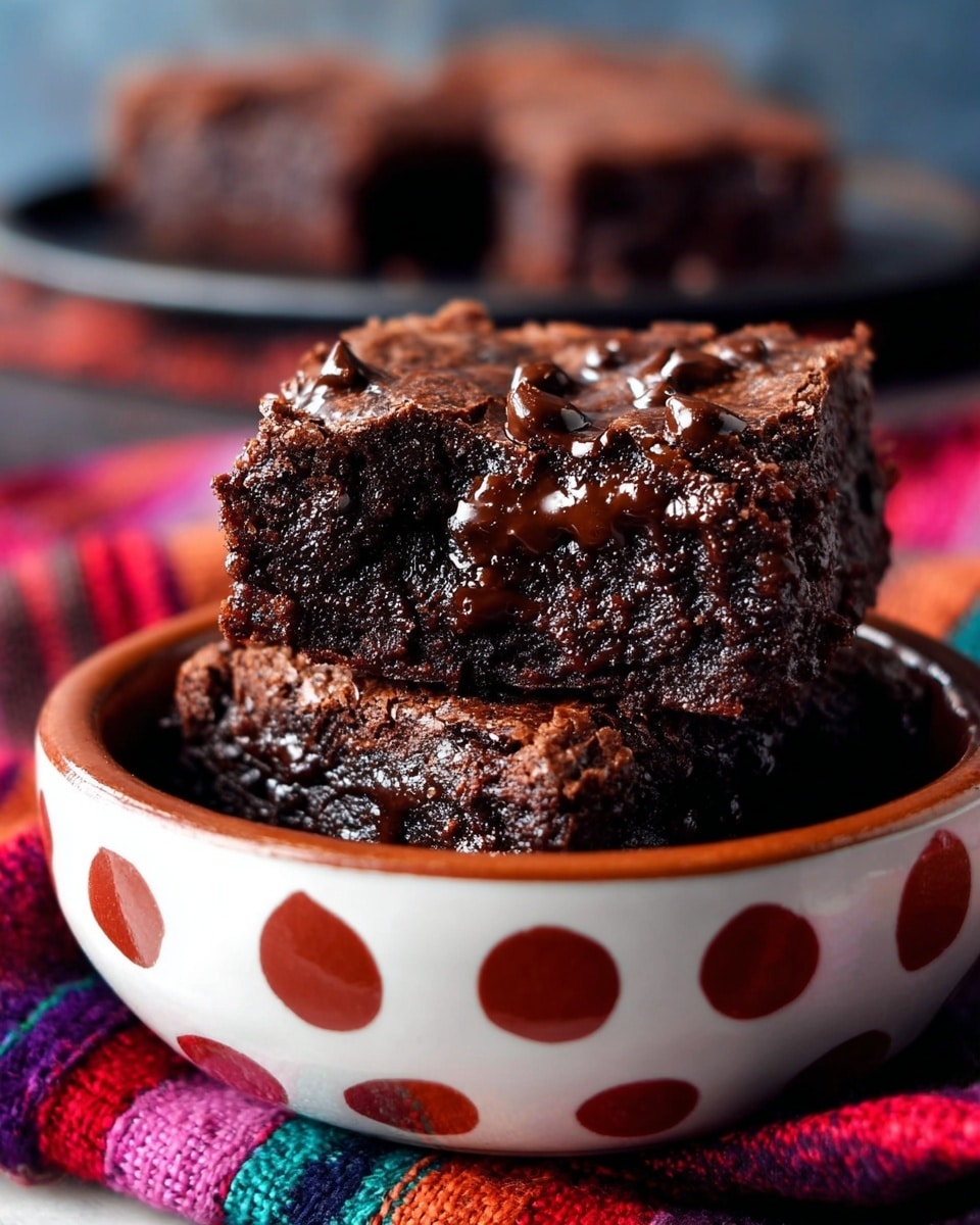 Two square pieces of rich dark brown chocolate brownies stacked inside a white bowl with red and white dot patterns. The brownies' top surface is slightly cracked with shiny, melted chocolate chips and a gooey dark filling visible between the layers. The bowl sits on a colorful striped fabric, with blurred dark chocolate brownies in the background on a dark round tray. The surface beneath all is a white marbled texture. photo taken with an iphone --ar 4:5 --v 7