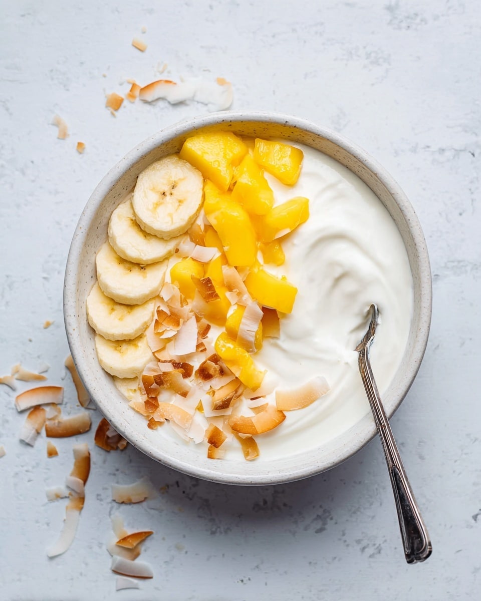 A white bowl filled with a smooth white yogurt base, topped with three separate sections of fruit and coconut flakes: round banana slices on the left, bright yellow mango chunks on the right, and toasted coconut flakes scattered mostly in the lower left. A metal spoon rests on the yogurt on the right side of the bowl. The bowl sits on a white marbled surface with a few coconut flakes scattered beside it. photo taken with an iphone --ar 4:5 --v 7