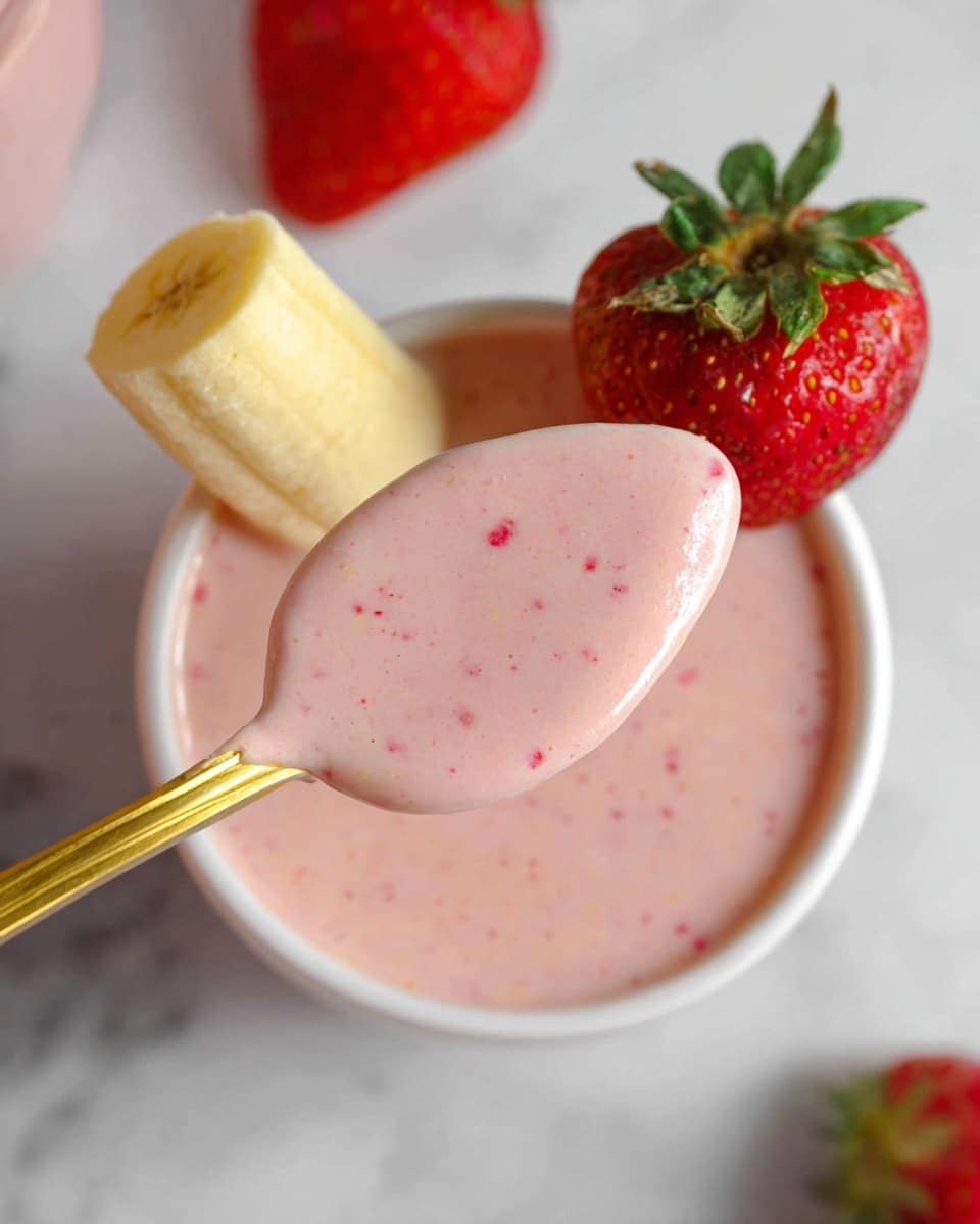 The image shows a white glass bowl filled with a smooth, pink strawberry yogurt that has small red specks throughout. On the edge of the bowl, there is a whole bright red strawberry with green leaves and a small piece of light yellow banana standing upright next to it. A golden spoon holds a portion of the yogurt close to the camera, showing the creamy, slightly thick texture and pink color with red specks. The setup is placed on a white marbled surface, and the focus centers on the yogurt spoon and the bowl below. photo taken with an iphone --ar 4:5 --v 7
